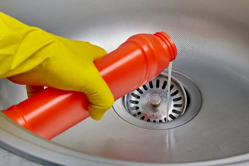 Worker's hand in a yellow rubber glove pouring liquid from a bottle down the metal sink's drain